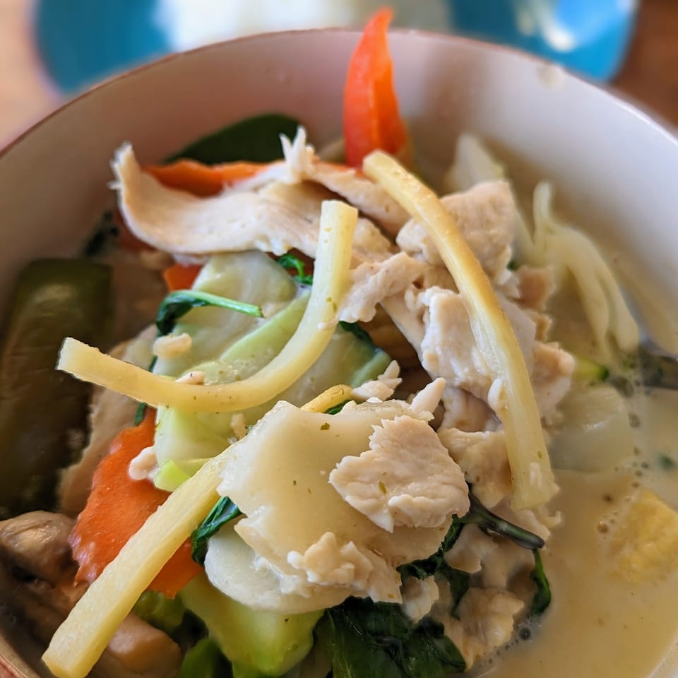 Close-up of a bowl of green curry with chicken, bamboo shoots, and mixed vegetables, with rice in the background.
