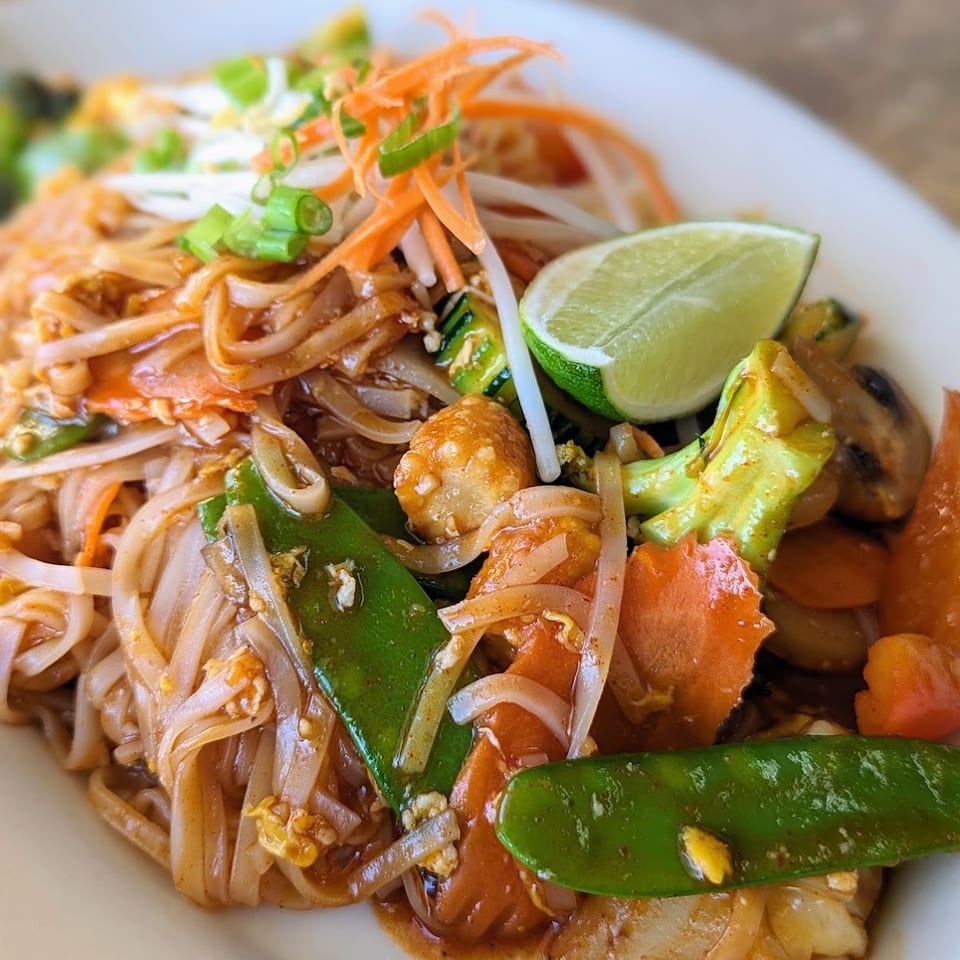 Close-up of Pad Thai noodles with vegetables, tofu, and a lime wedge on a white plate.